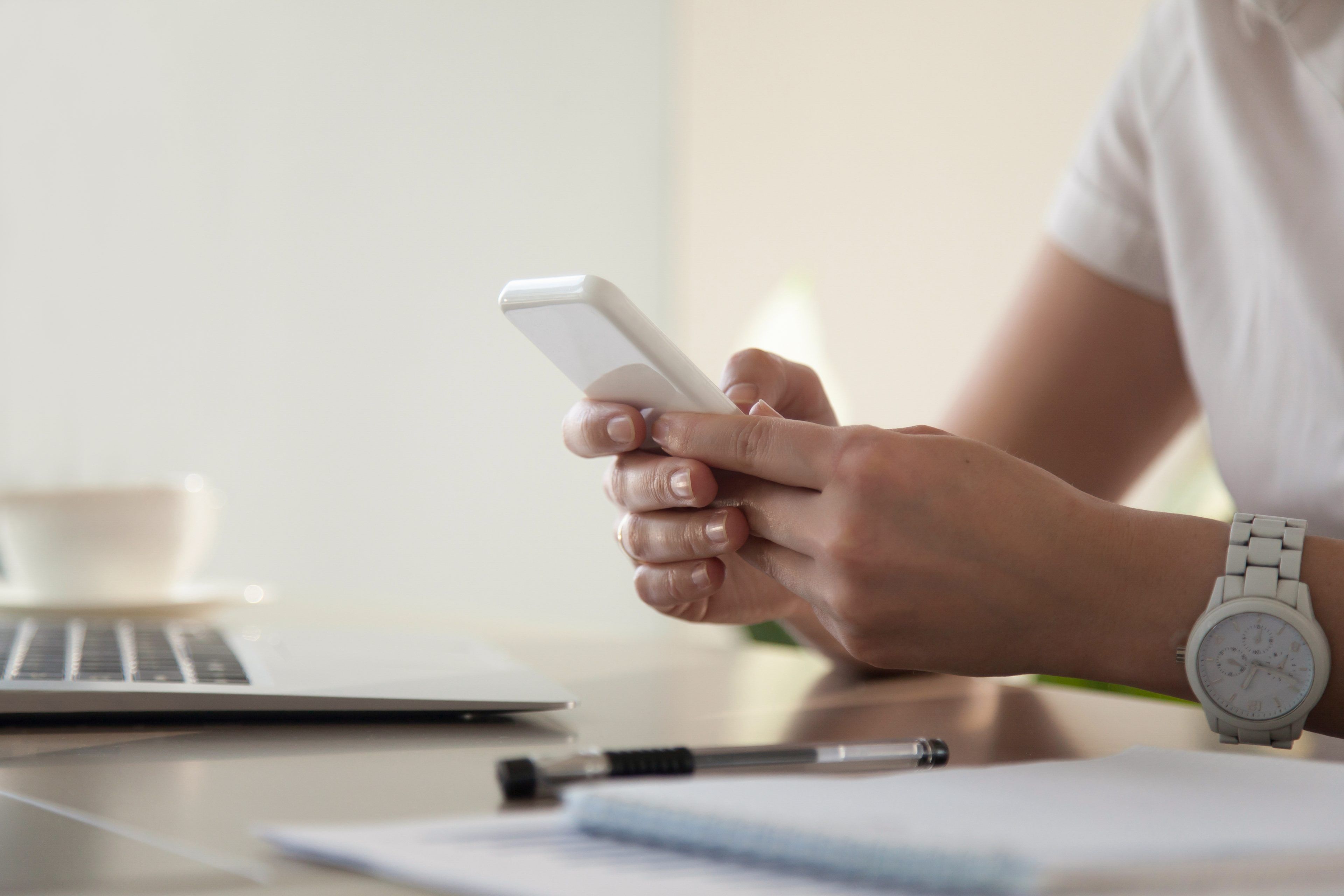 A person wearing a watch is using a smartphone at a desk with a laptop, notebook, and pen. The scene conveys focus and modern work life.