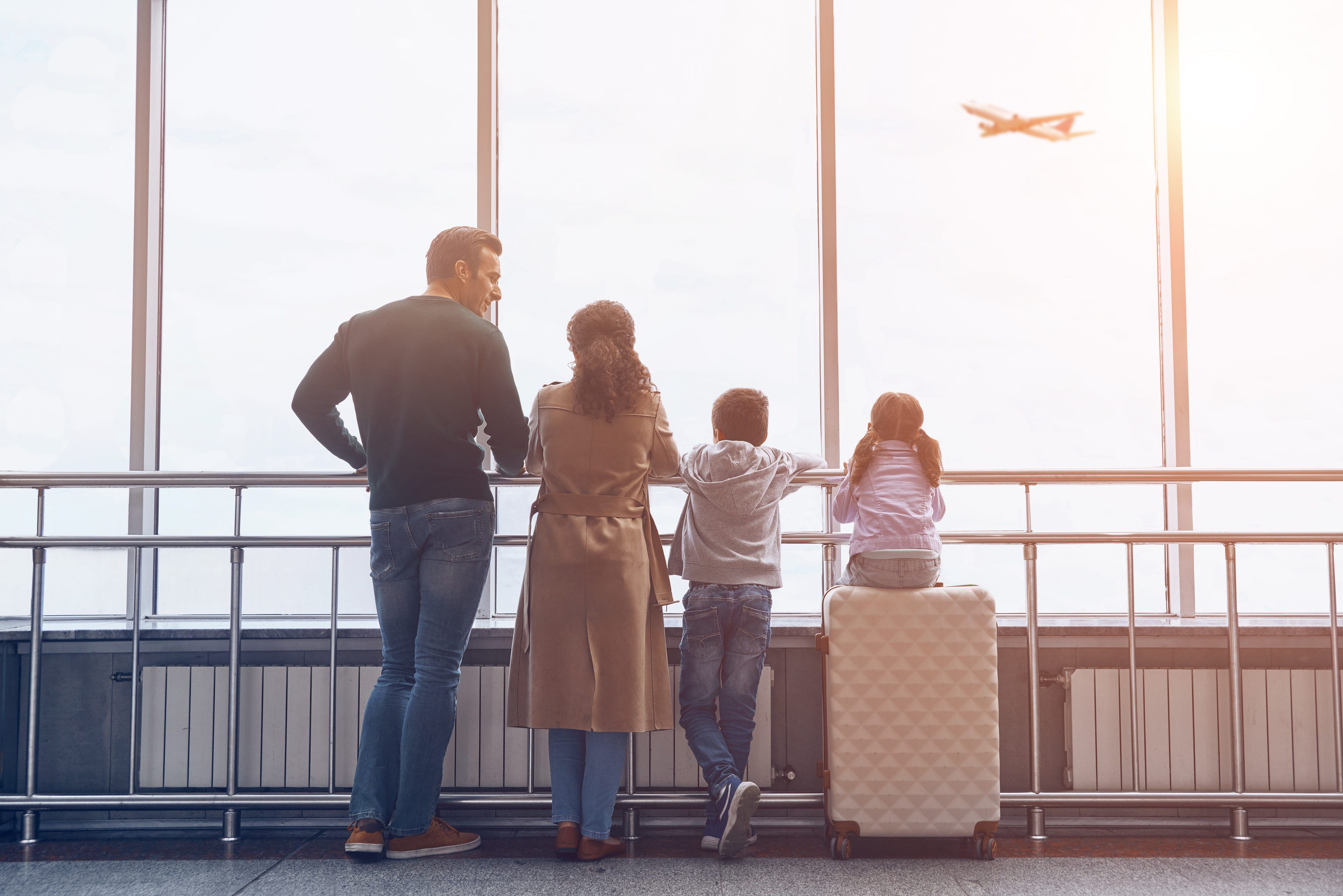 Family at an airport window, watching a plane take off. Two adults and two children stand together, with a suitcase beside them. The scene is warm and hopeful.