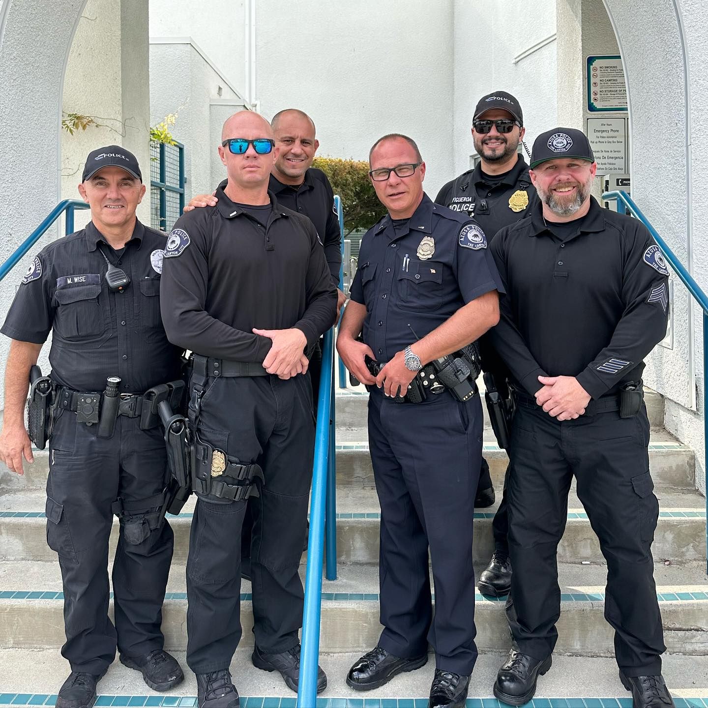 A group of six uniformed police officers stand together on outdoor steps, smiling. The scene is professional, friendly, and collaborative.